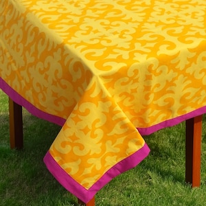 May include: Yellow tablecloth with a pink border and a repeating pattern of stylized swirls. The tablecloth is laid on a wooden table and grass.