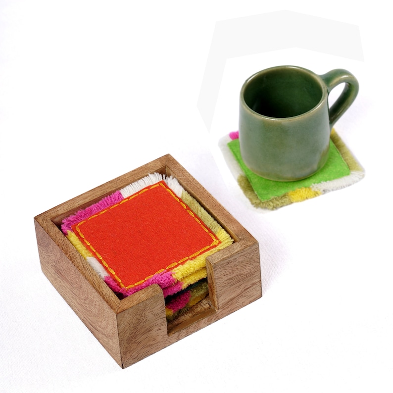 Puede incluir: Un juego de posavasos de tela coloridos con un soporte de madera. Los posavasos est&aacute;n apilados, con el posavasos superior que presenta un centro naranja brillante y costuras amarillas. El soporte es de madera natural y tiene una forma rectangular.