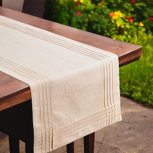 May include: A beige linen table runner with vertical pleats at the ends and along the sides. The runner is placed on a dark brown wooden table. The background features green grass and colorful flowers.