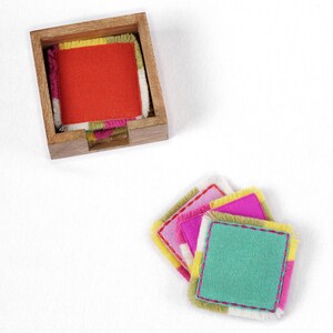 Puede incluir: Conjunto de posavasos de tela coloridos con bordes deshilachados. Los posavasos est&aacute;n apilados en un soporte de madera. Los posavasos son de color rosa, verde, amarillo y naranja.