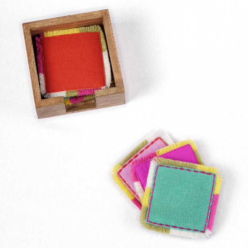 Puede incluir: Conjunto de posavasos de tela coloridos con bordes deshilachados. Los posavasos est&aacute;n apilados en un soporte de madera. Los posavasos son de color rosa, verde, amarillo y naranja.