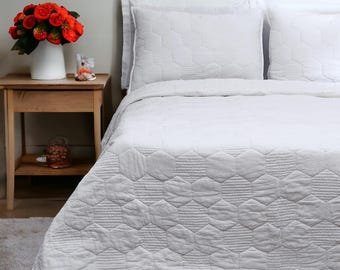 May include: A white bedspread and two matching pillow shams featuring a stitched hexagonal pattern. The bedding set is displayed on a bed next to a wooden nightstand holding a white vase filled with a bouquet of vibrant orange flowers.