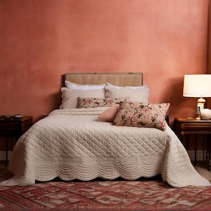 May include: A white quilted bedspread with a scalloped edge, on a bed with a woven headboard and a variety of patterned pillows. The bed is in a room with a terracotta colored wall and a wooden side table with a lamp.