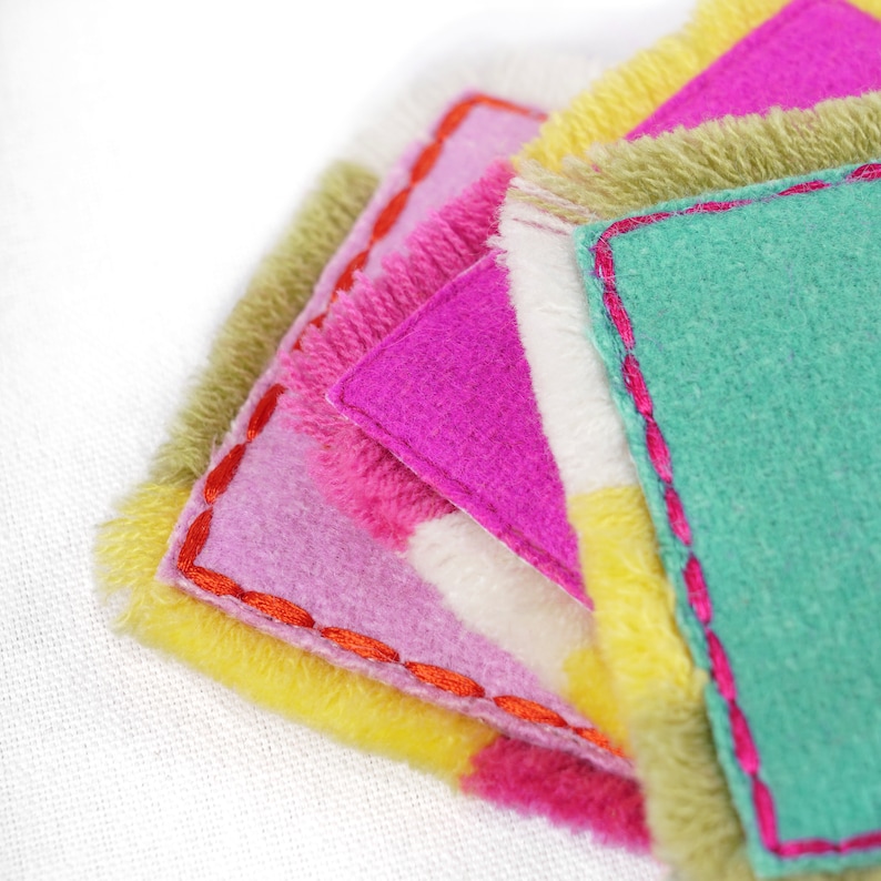 Puede incluir: Una pila de cuadrados de fieltro de colores con bordes deshilachados. Los cuadrados est&aacute;n superpuestos en un patr&oacute;n de arco&iacute;ris, con rosa, verde, amarillo y blanco.