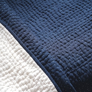 May include: Close-up of a navy blue and white quilted blanket. The navy blue section is on the top right, with a white section on the bottom left. The blanket has a textured, stitched pattern.