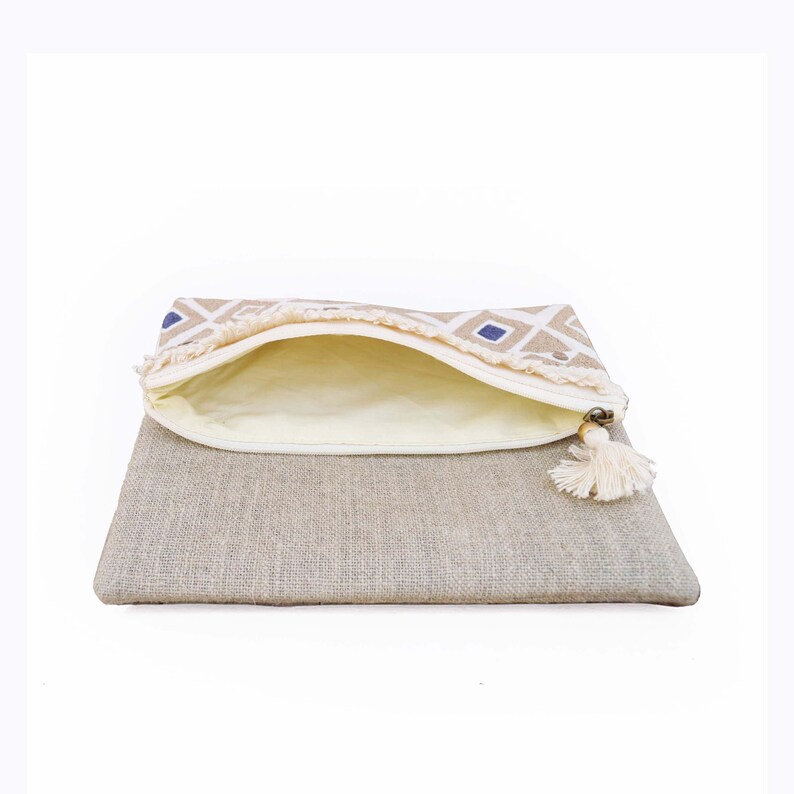 May include: A beige linen clutch bag with a folded top flap featuring a white and blue geometric diamond pattern. The bag has a cream-colored interior lining, a white zipper closure, and a decorative white tassel attached to the zipper pull.