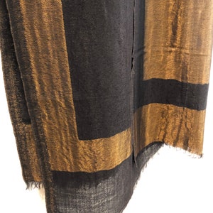 May include: A black and gold scarf with a geometric design. The scarf features wide gold stripes framing black rectangular sections. The fabric has a textured appearance and a fringed edge. The scarf is made of a soft material.