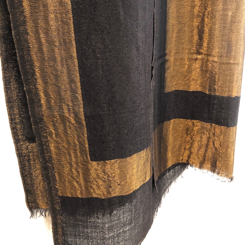 May include: A black and gold scarf with a geometric design. The scarf features wide gold stripes framing black rectangular sections. The fabric has a textured appearance and a fringed edge. The scarf is made of a soft material.