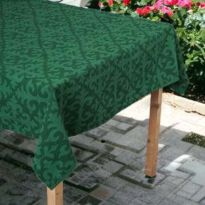 May include: A rectangular tablecloth in a deep forest green hue featuring a repeating dark green damask-style pattern. The fabric drapes over the edges of a wooden table, displaying a consistent geometric motif across the entire surface area.