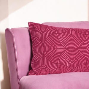 May include: A pink velvet sofa with a red cushion featuring a geometric pattern of white arches.