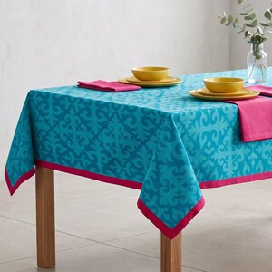 May include: A rectangular tablecloth in bright turquoise with a repeating dark blue damask-style pattern and a solid hot pink border. Two place settings are arranged on top, each featuring a yellow bowl on a matching yellow plate.