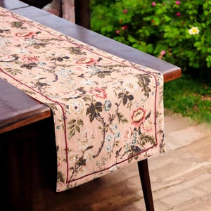 Könnte beinhalten: Ein rosa-farbener Tischläufer mit Blumenmuster und einem burgunderfarbenen Saum. Der Läufer liegt auf einem dunkelbraunen Holztisch.