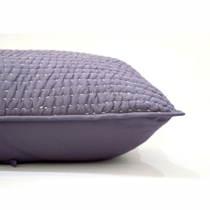 Puede incluir: Primer plano de una almohada acolchada de color morado con costuras blancas. La almohada se muestra de lado, con las costuras visibles en la parte superior y el borde de la almohada visible en la parte inferior.