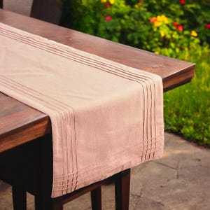 Op de afbeelding: Een lichtbruine tafelloper met een geplooid ontwerp. De loper is gemaakt van een zachte stof en is gedrapeerd over een donkerbruine houten tafel. De tafel staat buiten, met een vage achtergrond van groen gras en kleurrijke bloemen.