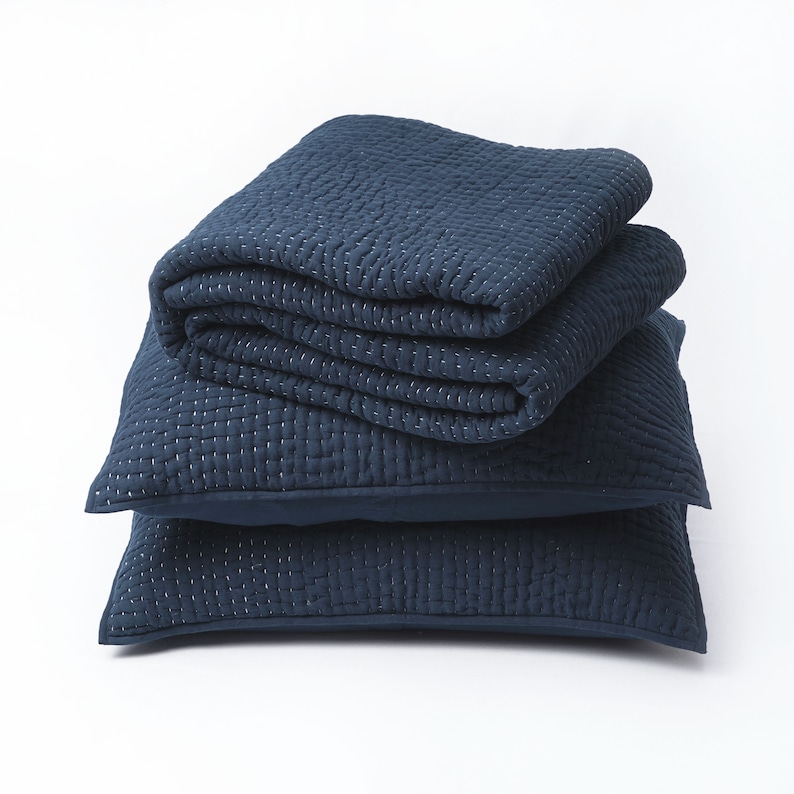 May include: A stack of navy blue bedding, including two square pillows and a folded blanket. The fabric has a quilted texture with white stitching. The items are arranged on a white background.