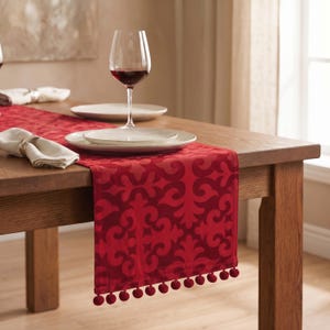 Puede incluir: Un camino de mesa rojo con un patrón de damasco tonal y una fila de pequeños pompones rojos en el borde inferior. El camino está colocado sobre una mesa de madera con platos blancos, una copa de vino tinto y servilletas de tela blancas.