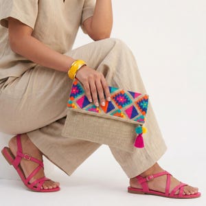 May include: A beige fold-over clutch bag featuring a vibrant geometric pattern in pink, orange, blue, and purple on the flap. The clutch includes a bright pink tassel accent attached to the side, paired with a yellow bangle and pink sandals.