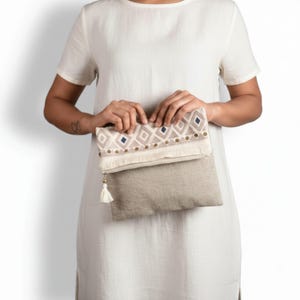 May include: A fold-over clutch bag featuring a beige linen lower section and a patterned upper flap. The flap displays a geometric diamond design in white, beige, and navy blue, accented with small gold studs, fringe trim, and a white tassel zipper pull.
