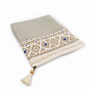 May include: A rectangular zippered pouch made of light beige linen fabric. The front features a decorative band with a geometric pattern in tan, white, and navy blue, finished with a fringe trim and a gold bead attached to the zipper pull.