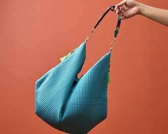 May include: A quilted turquoise hobo bag featuring a textured diagonal stitch pattern, gold-tone metal hardware, and a detachable shoulder strap with a colourful embroidered design. The bag is accented with bright lime green tassels on the sides.