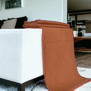 Puede incluir: Una manta de tejido de waffle marr&oacute;n drapeada sobre un sof&aacute; blanco. La manta est&aacute; doblada en la parte superior y cuelga hasta el suelo.