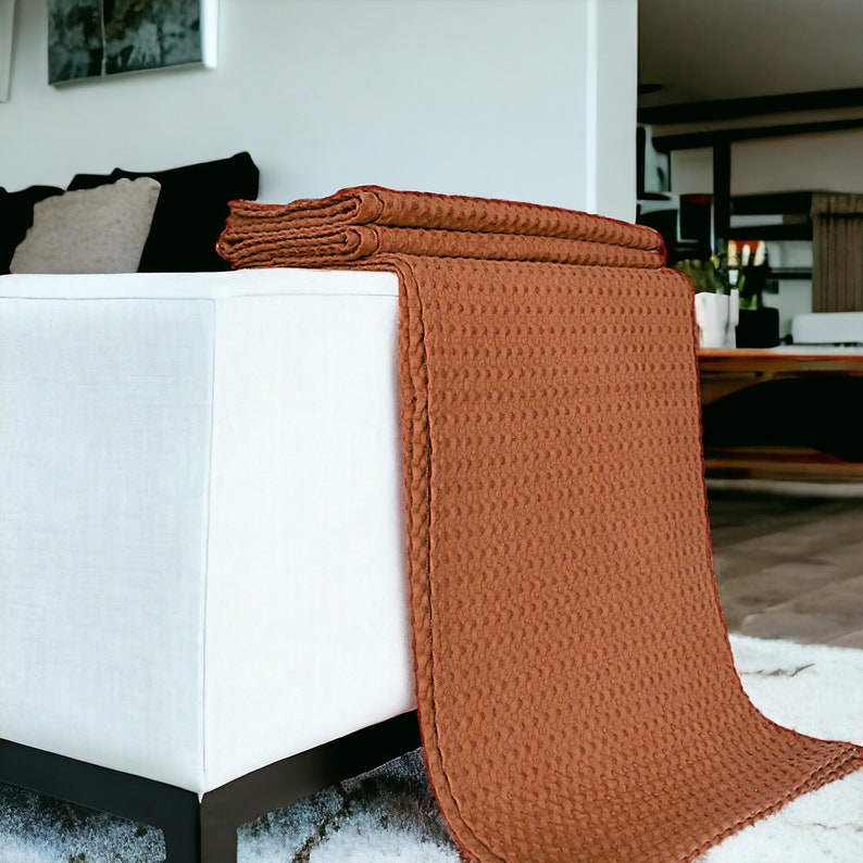 Puede incluir: Una manta de tejido de waffle marr&oacute;n drapeada sobre un sof&aacute; blanco. La manta est&aacute; doblada en la parte superior y cuelga hasta el suelo.