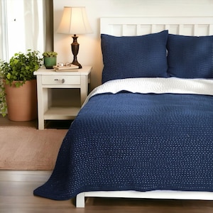 May include: A navy blue quilted bedspread and pillow set on a white bed frame. The bedspread features a dotted pattern. A white bedside table with a lamp and a potted plant are also visible.