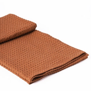 Puede incluir: Una manta de tejido de waffle marr&oacute;n doblada por la mitad. La manta est&aacute; hecha de un tejido suave y texturizado.