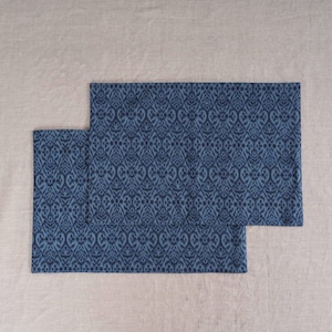 May include: Two blue placemats with a repeating, ornate pattern. The placemats are a darker shade of blue and are placed on a neutral-colored surface. The pattern is a stylized, symmetrical design.
