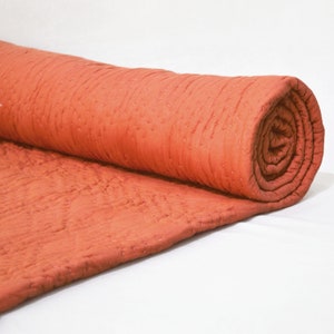 Op de afbeelding: Een opgerolde terracottakleurige quilt met een gestikt patroon. De quilt is gemaakt van een zachte, lichte stof.