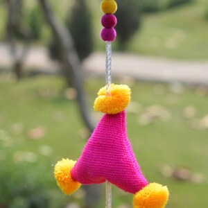 May include: A pink and yellow pom-pom garland with a triangular pink fabric piece in the center. The garland is hanging from a white string.