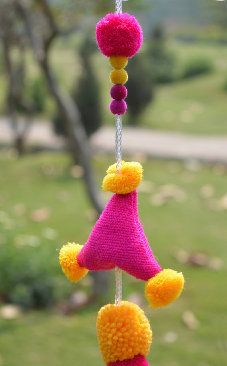 May include: A pink and yellow pom-pom garland with a triangular pink fabric piece in the center. The garland is hanging from a white string.