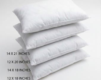 May include: A stack of four rectangular white pillows. The text on the left indicates sizes: 14 X 21 inches, 12 X 20 inches, 14 X 18 inches, and 12 X 18 inches. Each pillow has a plain white fabric cover and a soft, filled appearance.