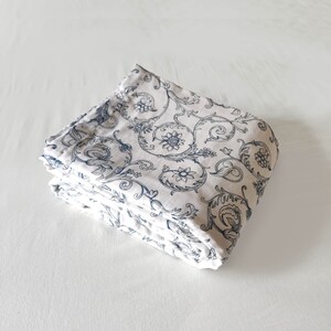 May include: A folded white fabric with a blue floral pattern. The fabric has a delicate, intricate design with swirling vines and small flowers.