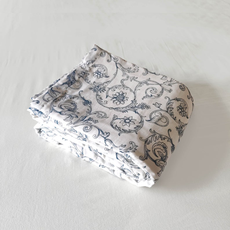 May include: A folded white fabric with a blue floral pattern. The fabric has a delicate, intricate design with swirling vines and small flowers.