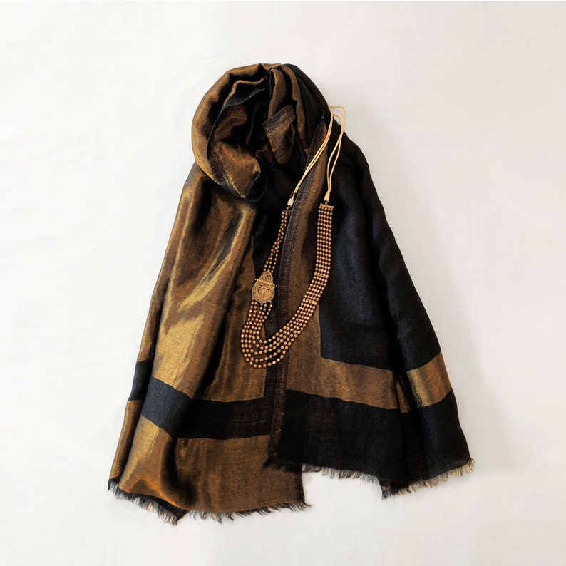 May include: A black and gold scarf with a geometric design and fringed edges. A multi-strand gold necklace with a pendant is draped over the scarf. The scarf has a metallic sheen.