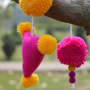 May include: A pink fabric heart with yellow pom-poms hanging from a branch. The heart is decorated with yellow pom-poms and has a string of yellow pom-poms hanging from the bottom.