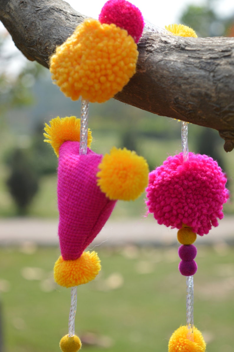 May include: A pink fabric heart with yellow pom-poms hanging from a branch. The heart is decorated with yellow pom-poms and has a string of yellow pom-poms hanging from the bottom.