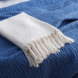 Könnte beinhalten: Eine weiße Decke mit Fransenrand. Die Decke ist gefaltet und auf einem blauen gesteppten Stoff gelegt.