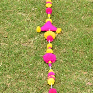 May include: A pink and yellow pom-pom garland with triangular shapes hangs on a green grassy lawn.