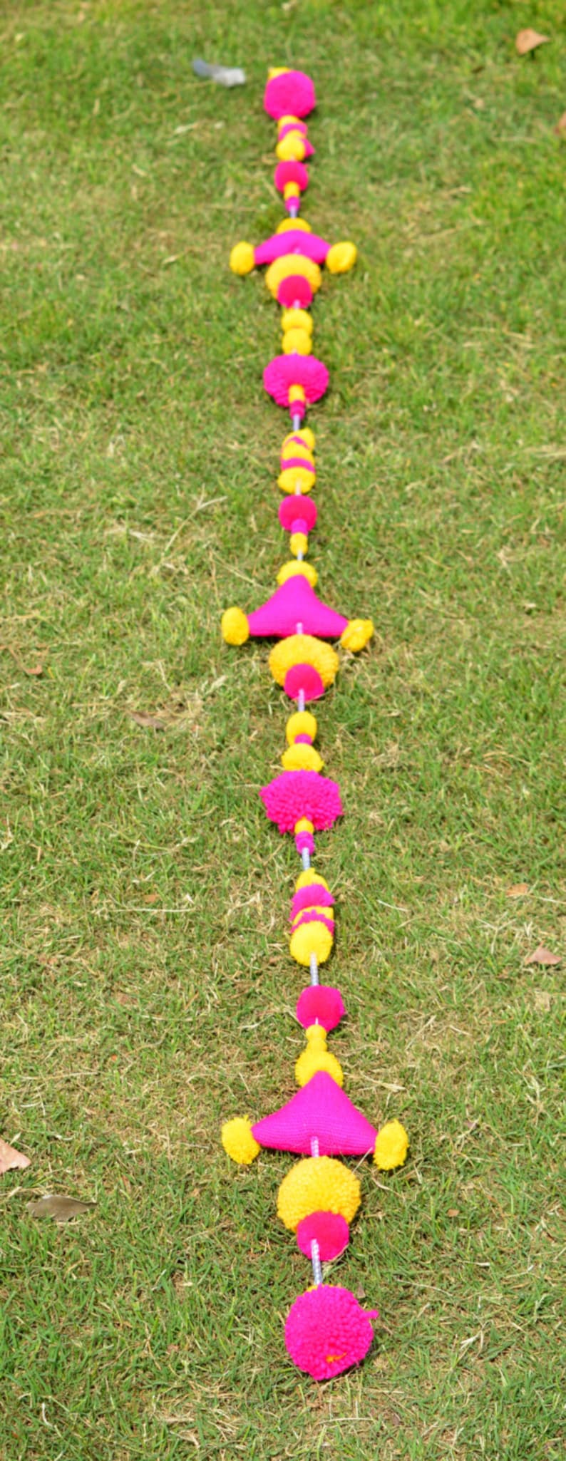 May include: A pink and yellow pom-pom garland with triangular shapes hangs on a green grassy lawn.
