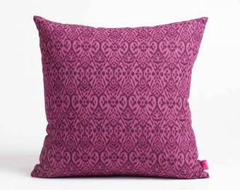 May include: A square decorative pillow in shades of purple. The pillow features a repeating pattern of stylized floral or paisley shapes. The fabric has a textured appearance. A small pink tag is attached to the bottom right corner of the pillow.