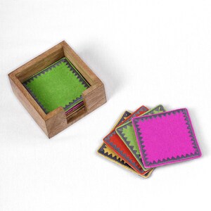Peut inclure: Un ensemble de six sous-verres color&eacute;s avec un bord festonn&eacute; gris. Les sous-verres sont empil&eacute;s et dispos&eacute;s dans un porte-sous-verres en bois.