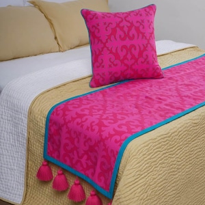 Op de afbeelding: Een decoratieve beddenset met een fuchsia kussen en loper met een herhalend patroon. De loper heeft een turquoise rand en roze kwastjes. Het bed is bedekt met een goudkleurig gewatteerd sprei en witte lakens.