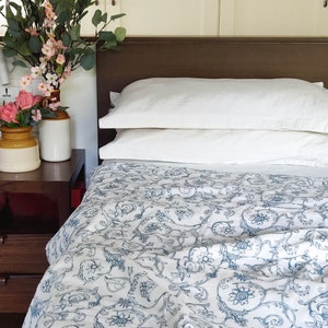 May include: A white bed with a blue and white floral patterned duvet cover and two white pillows. The bed is against a brown headboard and there are white cabinets in the background.