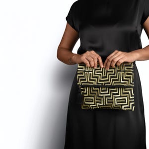 May include: A rectangular fold-over clutch bag featuring a black fabric base with an intricate, raised gold geometric maze pattern. The clutch includes a black zipper closure accented with a small black tassel hanging from the side zipper pull.