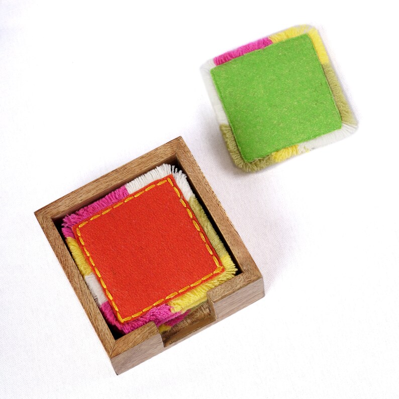 Puede incluir: Un juego de posavasos de tela coloridos con un patr&oacute;n amarillo, rosa, blanco y naranja. Los posavasos est&aacute;n en un soporte de madera con una ranura para una f&aacute;cil extracci&oacute;n. Los posavasos est&aacute;n hechos de fieltro y tienen un borde con flecos.