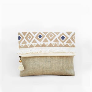May include: A rectangular clutch bag made of natural tan fabric. The top fold features a white band with embroidered tan diamonds, blue squares, and small circular accents. A white fringe trim lines the fold, and a white tassel hangs from the zipper.