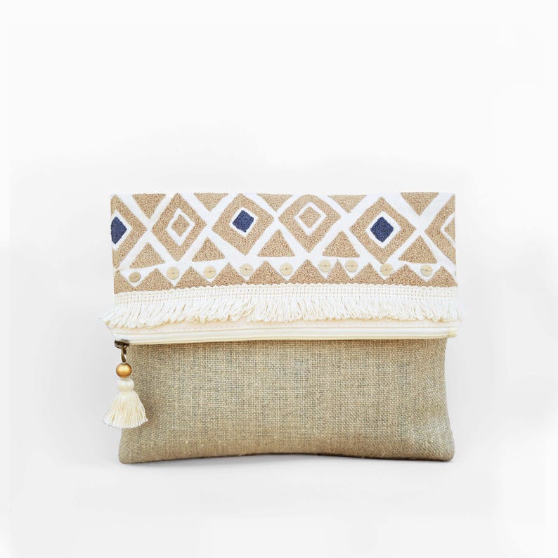 May include: A rectangular clutch bag made of natural tan fabric. The top fold features a white band with embroidered tan diamonds, blue squares, and small circular accents. A white fringe trim lines the fold, and a white tassel hangs from the zipper.