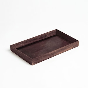 Puede incluir: Una bandeja rectangular de madera con acabado marrón oscuro. La bandeja tiene un diseño simple y minimalista con bordes elevados. La veta de la madera es visible, agregando textura a la superficie. La bandeja está sobre un fondo blanco.